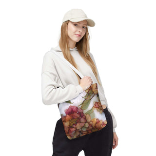 Woman wearing a beige hoodie and cap carrying a Vintage Peony Tote Bag with watercolor-style floral design in warm rose, apricot, and olive tones.